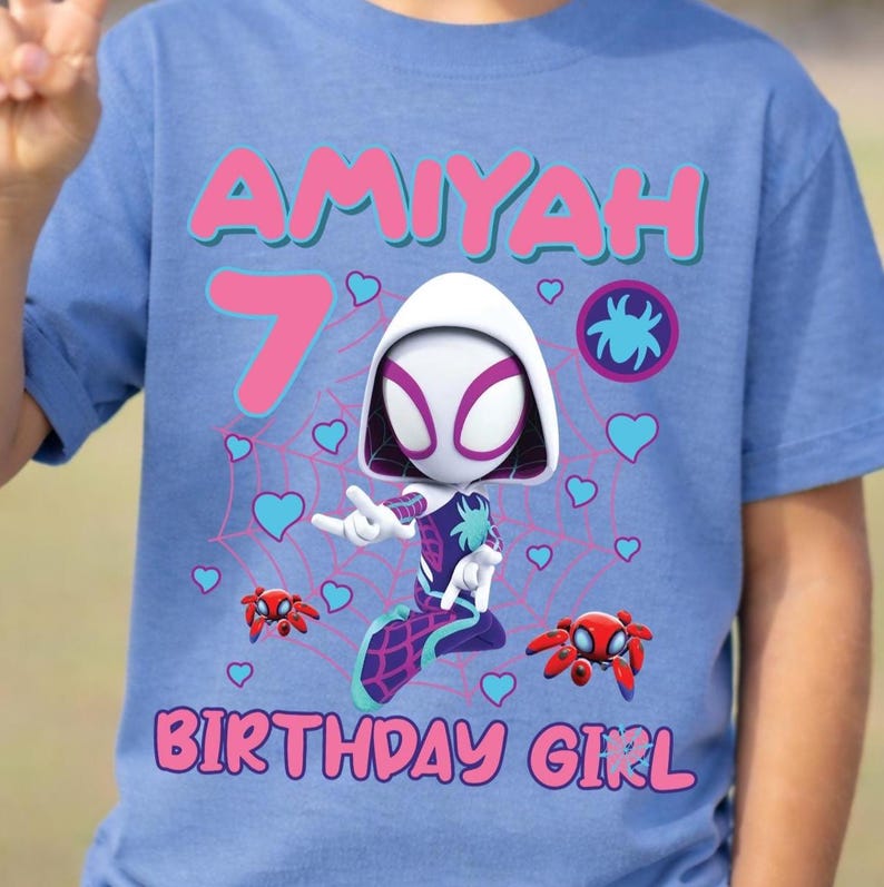 Puede incluir: Camiseta azul claro con un gr&aacute;fico de Spider-Gwen, el nombre "Amiyah" y el n&uacute;mero "7" en rosa. Tambi&eacute;n se muestran las palabras "BIRTHDAY GIRL". El dise&ntilde;o incluye corazones y elementos de telara&ntilde;a.