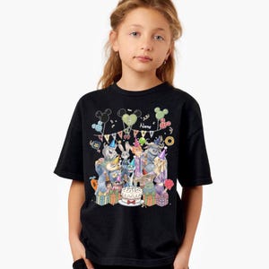Pu&ograve; includere: T-shirt nera con un design colorato per il compleanno con personaggi dei cartoni animati, palloncini e una torta. Il design include il numero "3" e la parola "Name" su uno striscione. La maglietta &egrave; indossata da una bambina.