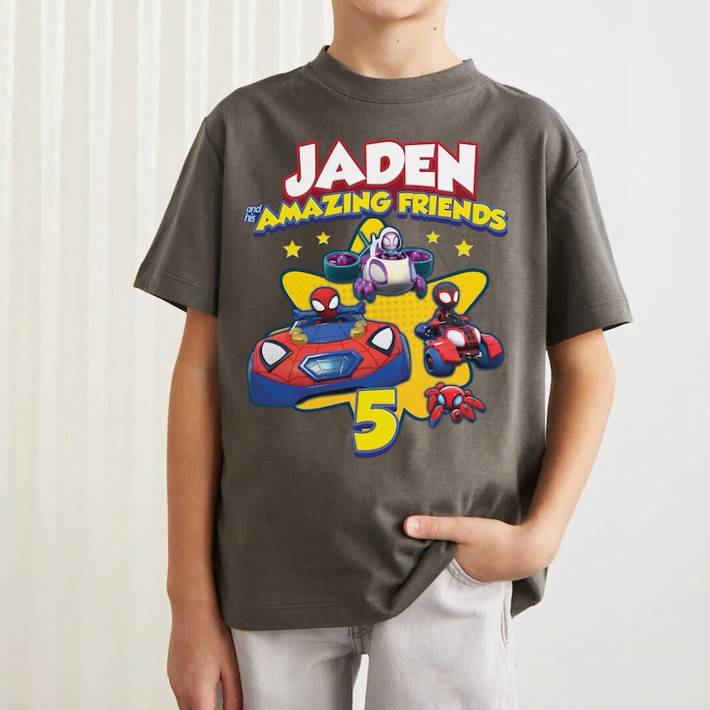 May include: A charcoal gray short-sleeve t-shirt featuring a graphic print with the text JADEN and his AMAZING FRIENDS. The design includes cartoon characters in vehicles, a large yellow star, and a prominent yellow number 5 on the front.