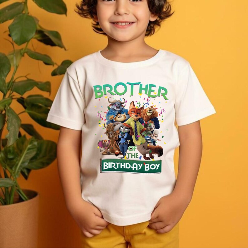 Pu&ograve; includere: Una maglietta bianca con la scritta "BROTHER OF THE BIRTHDAY BOY" in verde e bianco. La maglietta presenta una grafica colorata di personaggi dei cartoni animati. Il bambino indossa pantaloni gialli.