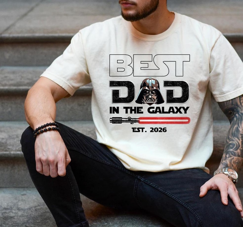 K&ouml;nnte beinhalten: Ein cremefarbenes Kurzarm-T-Shirt mit dem schwarzen Schriftzug BEST DAD IN THE GALAXY, einer Grafik von Darth Vaders Helm anstelle des Buchstabens A, einer roten Lichtschwert-Illustration und dem darunter in Schwarz gedruckten Text EST. 2026.