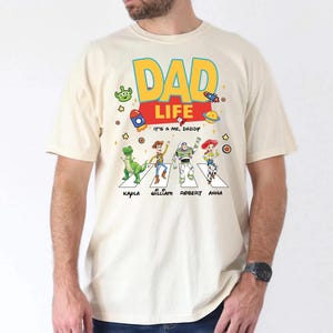 May include: Cream-colored t-shirt featuring a colorful graphic of Toy Story characters crossing a street, with the words "DAD LIFE" in large letters. Below the characters are the names Kapla, William, Robert, and Anna. The phrase "IT'S A ME, DADDY" is also present.