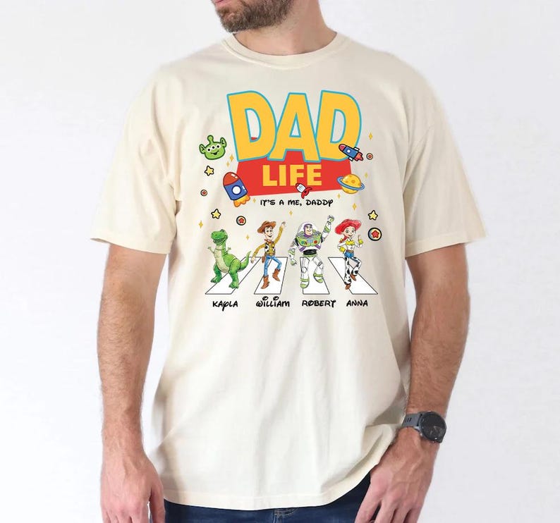 May include: Cream-colored t-shirt featuring a colorful graphic of Toy Story characters crossing a street, with the words "DAD LIFE" in large letters. Below the characters are the names Kapla, William, Robert, and Anna. The phrase "IT'S A ME, DADDY" is also present.