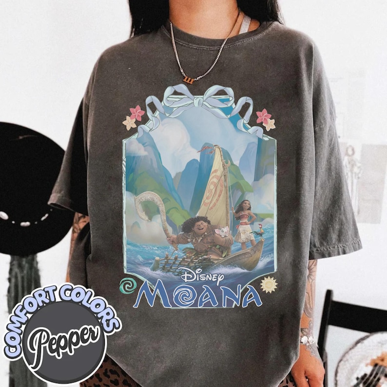 Puede incluir: Una camiseta de manga corta gris carb&oacute;n con un estampado gr&aacute;fico de Moana y Maui en un barco. El dise&ntilde;o incluye un lazo azul claro en la parte superior, flores tropicales y el texto Disney Moana. La camiseta tiene un acabado lavado vintage.