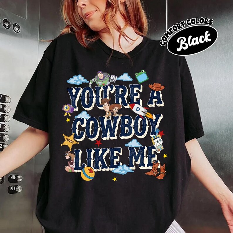 May include: A black short-sleeve t-shirt featuring the text "YOU'RE A COWBOY LIKE ME" in dark blue letters. The design includes various colorful cartoon characters and icons from a movie, such as a space ranger, a sheriff, a rocket, and boots.