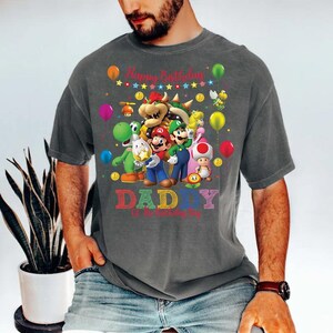 May include: Gray t-shirt featuring a colorful graphic of Mario and other characters from the Super Mario Bros. franchise. The shirt has the text "Happy Birthday DADDY" with balloons and stars. A man is wearing the shirt.