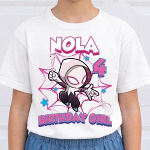 May include: White t-shirt featuring a cartoon illustration of a superhero character. The design includes the name "NOLA", the number "4", and the text "BIRTHDAY GIRL" in pink and blue. The character is set against a pink spiderweb background.
