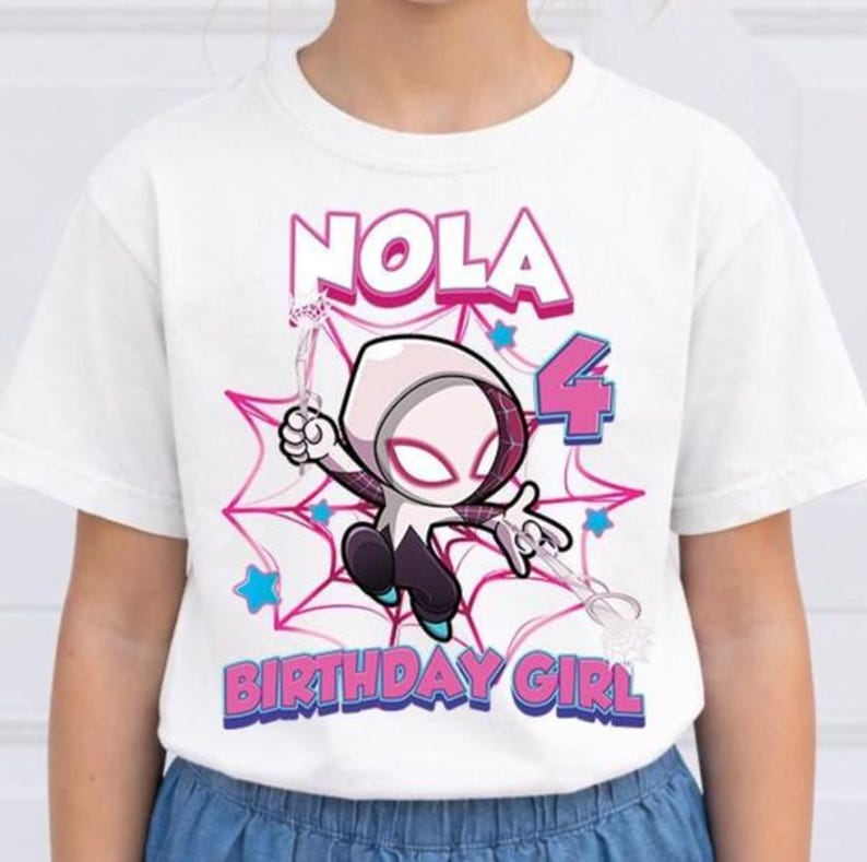 May include: White t-shirt featuring a cartoon illustration of a superhero character. The design includes the name "NOLA", the number "4", and the text "BIRTHDAY GIRL" in pink and blue. The character is set against a pink spiderweb background.