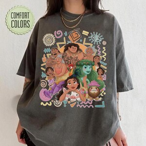 May include: A charcoal gray short-sleeve t-shirt featuring a colorful graphic print of various animated characters from the movie Moana, including Moana, Maui, and Heihei, surrounded by abstract shapes and the text Comfort Colors in the top corner.