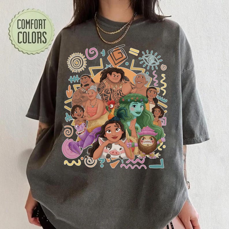 May include: A charcoal gray short-sleeve t-shirt featuring a colorful graphic print of various animated characters from the movie Moana, including Moana, Maui, and Heihei, surrounded by abstract shapes and the text Comfort Colors in the top corner.