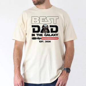 K&ouml;nnte beinhalten: Ein cremefarbenes Kurzarm-T-Shirt mit dem Text BEST DAD IN THE GALAXY, wobei der Helm von Darth Vader den Buchstaben A ersetzt. Unter dem Text befindet sich eine Grafik eines roten Lichtschwerts und der Schriftzug EST. 2026 in Schwarz.