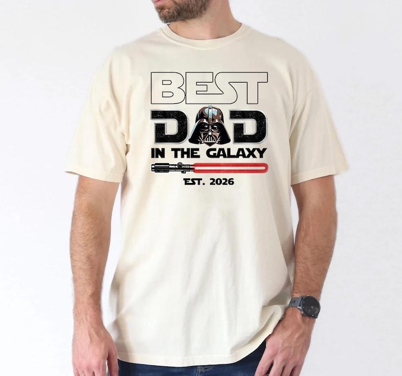 K&ouml;nnte beinhalten: Ein cremefarbenes Kurzarm-T-Shirt mit dem Text BEST DAD IN THE GALAXY, wobei der Helm von Darth Vader den Buchstaben A ersetzt. Unter dem Text befindet sich eine Grafik eines roten Lichtschwerts und der Schriftzug EST. 2026 in Schwarz.