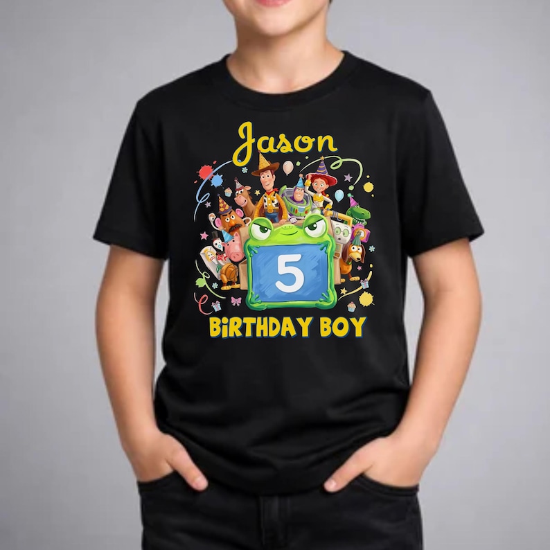 Pu&ograve; includere: T-shirt nera con grafica colorata con la scritta "Jason" e "Birthday Boy". La grafica include personaggi dei cartoni animati e il numero 5 su sfondo blu. La maglietta &egrave; indossata da una persona.