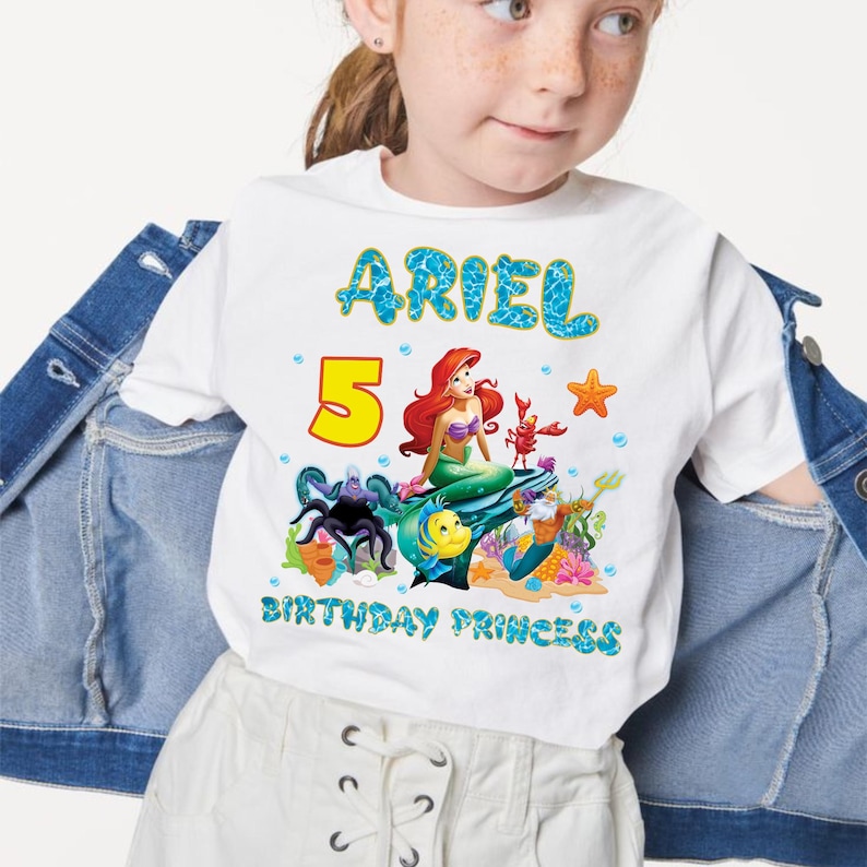 May include: White t-shirt featuring a colorful graphic of Ariel from The Little Mermaid, with the name "ARIEL" in blue bubble letters. The number "5" is in yellow, and the text "BIRTHDAY PRINCESS" is in blue.