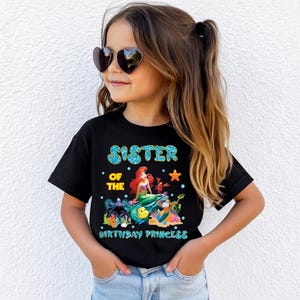May include: Black t-shirt with the text "SISTER OF THE BIRTHDAY PRINCESS" in blue and yellow. The shirt features a graphic of Ariel from The Little Mermaid with other characters. The girl is wearing heart-shaped sunglasses.