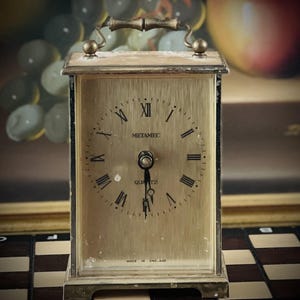 Puede incluir: Un reloj de cuarzo Metamec vintage con una esfera rectangular dorada y números romanos. El reloj tiene un asa y está marcado "Made in England". El reloj está sobre una superficie a cuadros.