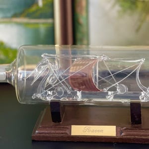 Puede incluir: Un barco de cristal transparente dentro de una botella. El barco tiene una vela roja y está montado sobre un soporte de madera con una placa dorada que dice "Roaven". La botella tiene un tapón redondo.