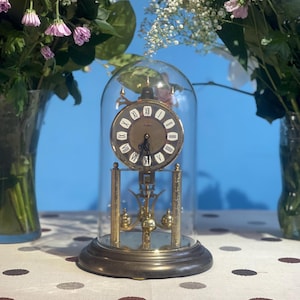 Può includere: Un orologio vintage in ottone con cupola di vetro, con un quadrante rotondo con numeri romani. L'orologio ha un pendolo ed è posto su una base scura. Lo sfondo include vasi di fiori rosa e bianchi e una tovaglia a pois.