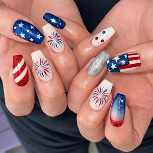 May include: A set of coffin-shaped acrylic nails with a patriotic theme. Designs include blue nails with white stars, red and white stripes, fireworks, and a silver glitter nail. The color scheme is red, white, and blue.
