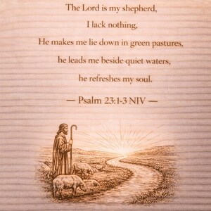 Op de afbeelding: Houten plaat met gegraveerde tekst uit Psalm 23:1-3 NIV. Het kunstwerk toont een herder met schapen, een rivier en een zonsopgang. De tekst luidt: "De Heer is mijn herder, mij ontbreekt het aan niets..." in een schreefletter.