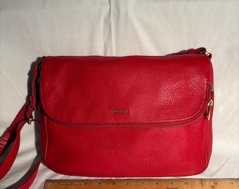 May include: A red leather crossbody bag with a flap closure. The bag has a textured surface and a long adjustable strap. The brand name "FOSSIL" is visible on the front. A ruler is placed below the bag.