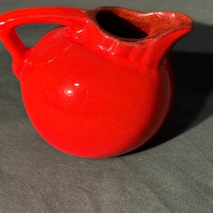 May include: A small, round, red ceramic pitcher with a handle and a spout. The pitcher has a glossy finish and a dark interior. The handle is curved, and the spout is slightly flared. The pitcher is likely for serving liquids.