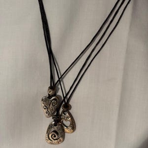 May include: Three pendant necklaces with black cords. The pendants are silver and gold-toned, featuring heart, teardrop, and oval shapes. Each pendant has decorative patterns.