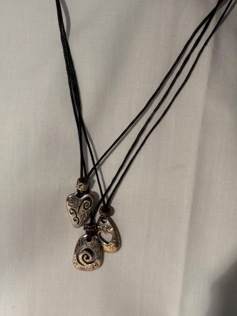 May include: Three pendant necklaces with black cords. The pendants are silver and gold-toned, featuring heart, teardrop, and oval shapes. Each pendant has decorative patterns.