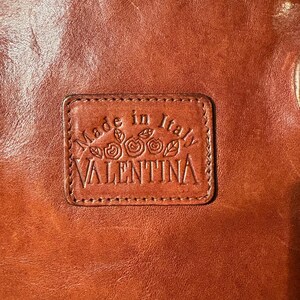 Peut inclure: Gros plan sur un article en cuir marron avec un patch rectangulaire en relief. Le patch indique "Made in Italy" au-dessus du nom "VALENTINA" avec des motifs floraux. Le cuir a une finition brillante.