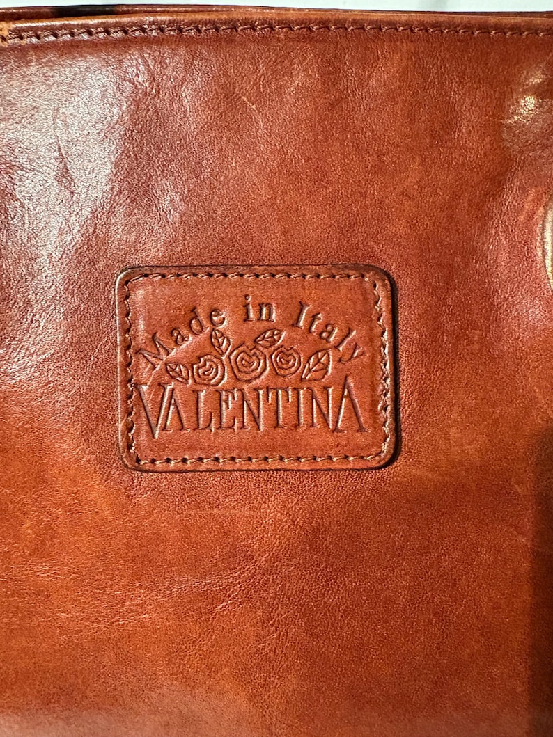 Peut inclure: Gros plan sur un article en cuir marron avec un patch rectangulaire en relief. Le patch indique "Made in Italy" au-dessus du nom "VALENTINA" avec des motifs floraux. Le cuir a une finition brillante.
