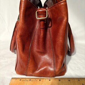 Peut inclure: Un sac fourre-tout en cuir marron de forme rectangulaire avec une poign&eacute;e. Le sac a une boucle dor&eacute;e et une r&egrave;gle est plac&eacute;e au bas du sac. Le sac mesure environ 18 cm de large.