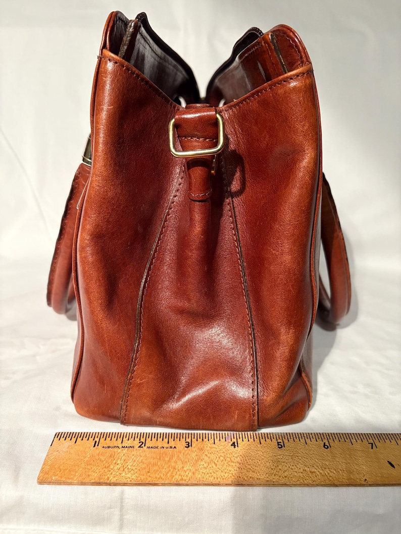 Peut inclure: Un sac fourre-tout en cuir marron de forme rectangulaire avec une poign&eacute;e. Le sac a une boucle dor&eacute;e et une r&egrave;gle est plac&eacute;e au bas du sac. Le sac mesure environ 18 cm de large.
