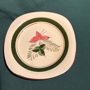 May include: A round, off-white ceramic plate with a hand-painted floral design. The plate features a central pink flower with green leaves and a green border. The rim is a reddish-brown color.