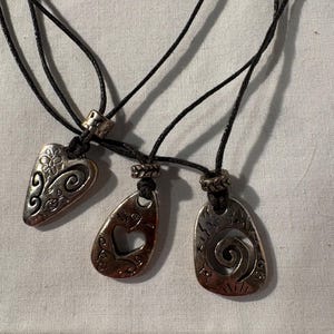 May include: Three silver-coloured pendant necklaces with black cords. Each pendant has a unique design: a floral heart, a teardrop with a heart cutout, and a spiral. The pendants are displayed on a white surface.