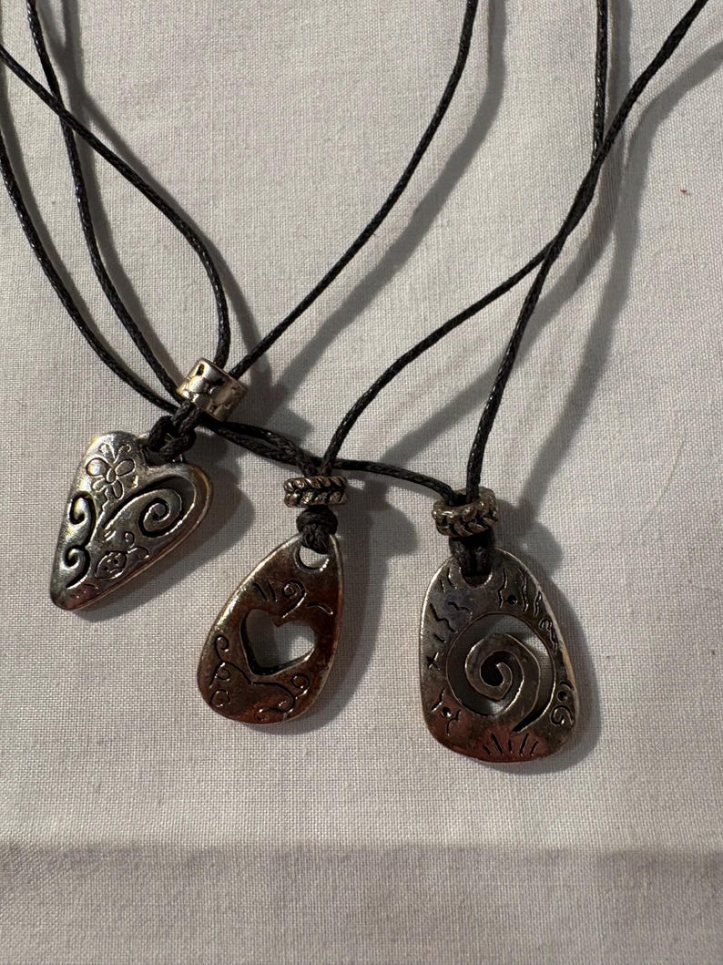 May include: Three silver-coloured pendant necklaces with black cords. Each pendant has a unique design: a floral heart, a teardrop with a heart cutout, and a spiral. The pendants are displayed on a white surface.