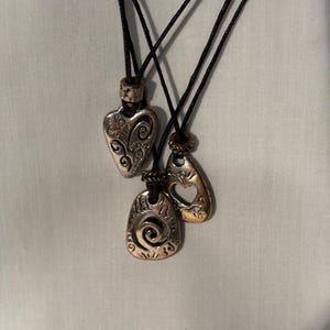 May include: Three silver-coloured pendant necklaces with black cords. The pendants have different shapes: a heart with floral details, a teardrop with a cutout, and a rectangular shape with a spiral design. The necklaces are displayed against a white background.