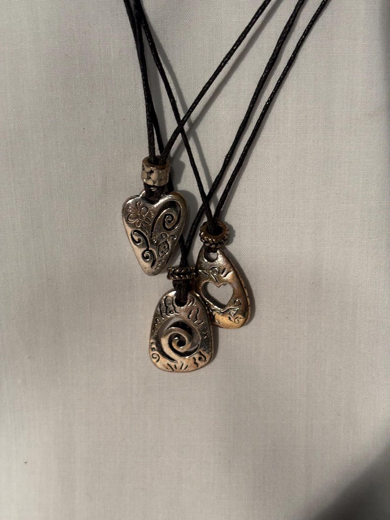 May include: Three silver-coloured pendant necklaces with black cords. The pendants have different shapes: a heart with floral details, a teardrop with a cutout, and a rectangular shape with a spiral design. The necklaces are displayed against a white background.