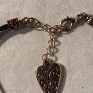 May include: Close-up of a silver heart pendant necklace. The pendant features a floral and swirl design. The necklace has a black cord and a gold-coloured chain with a lobster clasp.