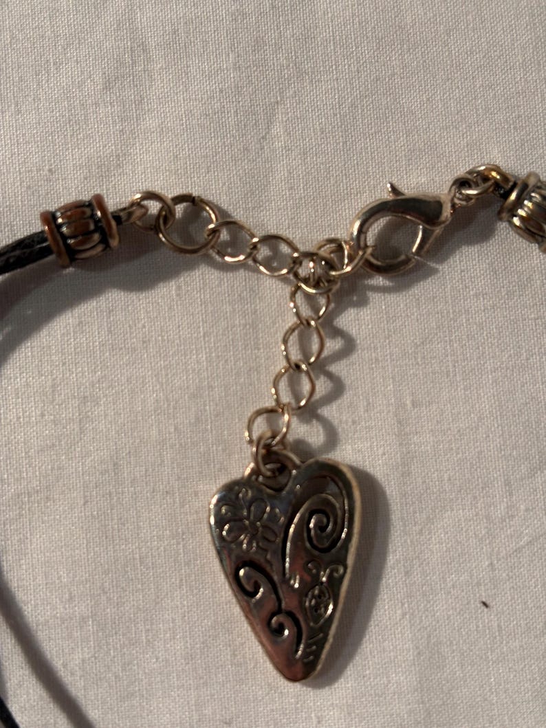 May include: Close-up of a silver heart pendant necklace. The pendant features a floral and swirl design. The necklace has a black cord and a gold-coloured chain with a lobster clasp.