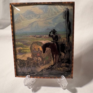 May include: A framed artwork depicting a Western scene with a cowboy on horseback, a covered wagon, and a desert landscape. The artwork has a brown wooden frame and is displayed on a clear stand. The background features mountains and a cloudy sky.