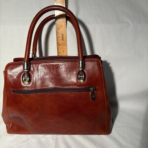 Peut inclure: Un sac &agrave; main en cuir marron avec deux poign&eacute;es sup&eacute;rieures et une poche zipp&eacute;e &agrave; l'avant. Le sac a une quincaillerie de couleur argent&eacute;e et une forme rectangulaire. Le cuir a une teinte riche et chaleureuse.