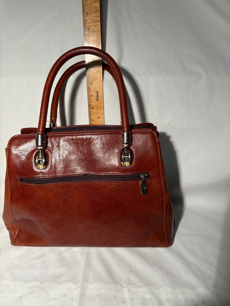 Peut inclure: Un sac &agrave; main en cuir marron avec deux poign&eacute;es sup&eacute;rieures et une poche zipp&eacute;e &agrave; l'avant. Le sac a une quincaillerie de couleur argent&eacute;e et une forme rectangulaire. Le cuir a une teinte riche et chaleureuse.