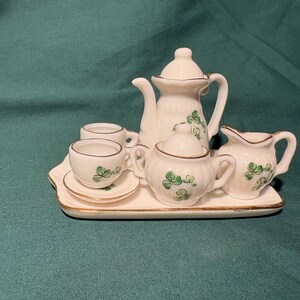 May include: A miniature white ceramic tea set with green shamrock designs. The set includes a teapot, creamer, sugar bowl, two teacups, and saucers, all arranged on a rectangular tray with gold trim. The tea set is a charming decorative item.