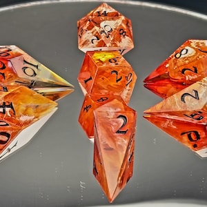 Puede incluir: Un conjunto de dados de resina naranja y transparente para juegos de mesa. Los dados tienen varias formas, incluyendo un d20, d12, d10, d8 y d6. Los números están pintados de negro. Los dados están dispuestos sobre una superficie reflectante.