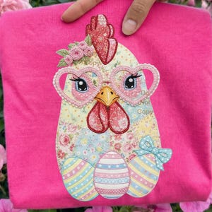 May include: A pink sweatshirt with an embroidered chicken design. The chicken wears heart-shaped glasses and a floral headpiece. Below the chicken are three decorated Easter eggs. The words "Do Not Copy" are visible.