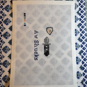 May include: Needlepoint canvas with a design featuring a shield, a black and gray object, and the text "Av Shucks" in navy blue. The canvas also includes a color palette on the upper left corner.