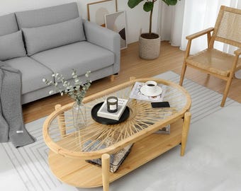 May include: A light wood oval coffee table with a glass top and woven rattan detailing. The table has a lower shelf and is surrounded by a gray sofa, a woven chair, and a rug. A candle, book, and coffee cup are on the table.