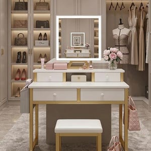 May include: A white vanity table with gold accents, a lighted mirror, and a matching stool. The vanity has drawers and a shelf for storage. A closet with handbags, shoes, and dresses is in the background. A bouquet of pink flowers sits on the vanity.