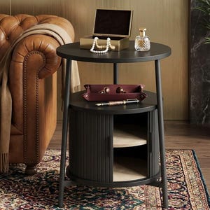 May include: A black, two-tiered side table with a round top and a storage cabinet. The top shelf holds a gold jewelry box, a perfume bottle, and a pearl necklace. The lower shelf has a leather tray with cufflinks and a pen.