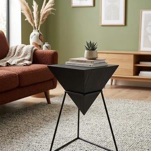 May include: A black geometric side table with a triangular top and a black metal frame. The table is holding a small potted plant and a stack of books. The room has a neutral color palette with a brown sofa and a light gray rug.